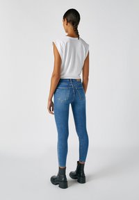 Jeans ajustados de tiro alto en azul con bolsillos traseros, combinados con una blusa blanca de manga corta y botas de tobillo negras de suela gruesa. Textura de denim suave.