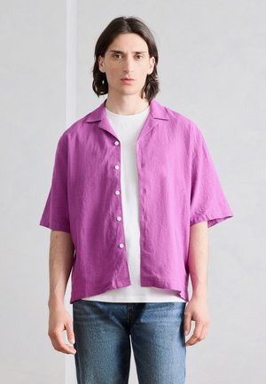 Νεαρό άτομο με σκούρα μαλλιά μέχρι τους ώμους, που φοράει φωτεινό μωβ κοντομάνικο πουκάμισο πάνω από άσπρο μπλουζάκι και μπλε τζιν.