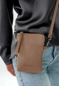 Hnědá kožená crossbody taška s texturovaným povrchem, opatřená zipem a odnímatelným ramenním popruhem, vystavená na tmavé blůze.
