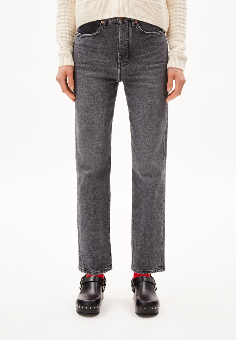 Personne portant un jean droit gris, un pull crème, des chaussettes rouges et des sabots noirs cloutés, debout sur un fond blanc uni.