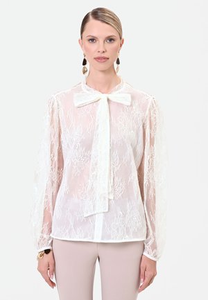 Donna che indossa una camicetta bianca in pizzo trasparente con un grande fiocco al colletto e maniche lunghe, abbinata a pantaloni beige chiaro.