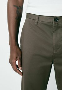 Pantaloni din material texturat, de culoare maro, cu față plată, buzunare standard și închidere cu un singur nasture. Se vede o mână odihnindu-se pe lateral.