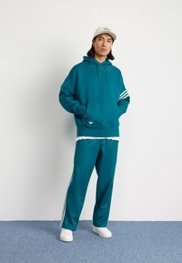 Teal hettegenser med frontlomme og hvite striper på ermene, parret med teal joggebukser med sidelister, båret med hvite sneakers.