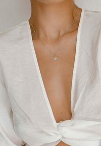 Collar de plata con un colgante floral, llevado con una blusa blanca de textura y escote profundo en forma de V atada en la cintura. Piel visible en el escote.