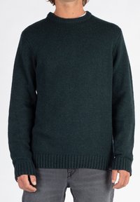 Man in een donkergroene ribgebreide trui en grijze broek, staande tegen een effen lichte achtergrond.