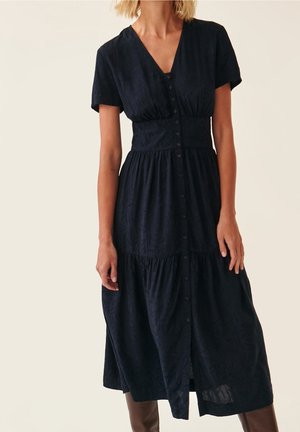 Femme portant une robe midi noire à manches courtes, boutonnée, taille cintrée et motif texturé subtil.