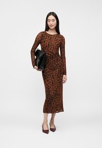 Naine kannab pruuni ja musta loomamustriga pikkade varrukatega kleiti, hoides musta käekotti ja jalas burgundia punased terava ninaga kingad.