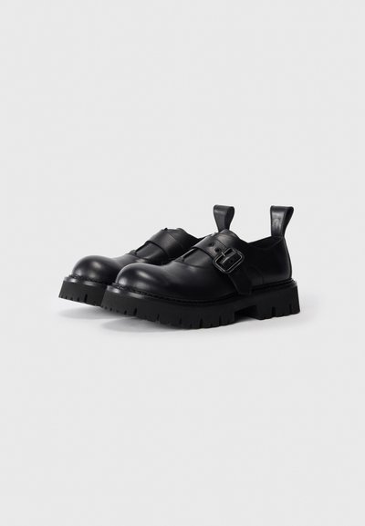 Pantofi din piele neagră cu o talpă groasă și texturată. Dotate cu o cataramă și o curea pentru gleznă. Design cu vârf rotund. Stil minimalist cu cârlige pentru a fi îmbrăcați.