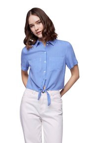 Camicia a maniche corte a righe blu e bianche con colletto, dotata di un dettaglio con nodo frontale. Indossata con pantaloni chiari.