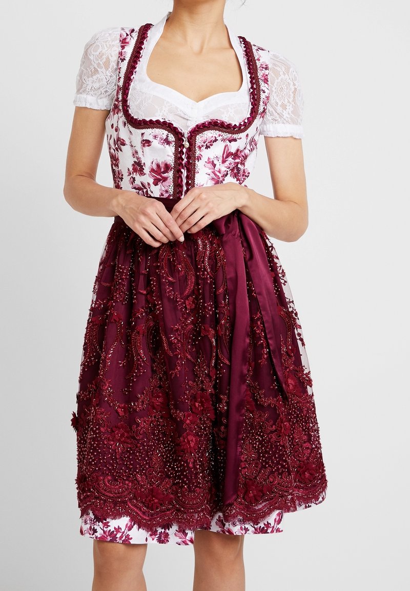 Femme portant une robe dirndl fleurie blanche et bordeaux avec des manches en dentelle et un tablier bordeaux brodé noué à la taille.