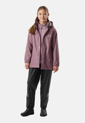 Młoda dziewczyna w mauve kapturze przeciwdeszczowej kurtce, czarnych wodoodpornych spodniach i czarnych butach, stojąca przodem na białym tle.