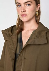 Braune Kapuzenjacke mit einem Reißverschluss, Druckknöpfen und matter Textur, getragen über einem karierten Hemd mit Blau- und Grüntönen.