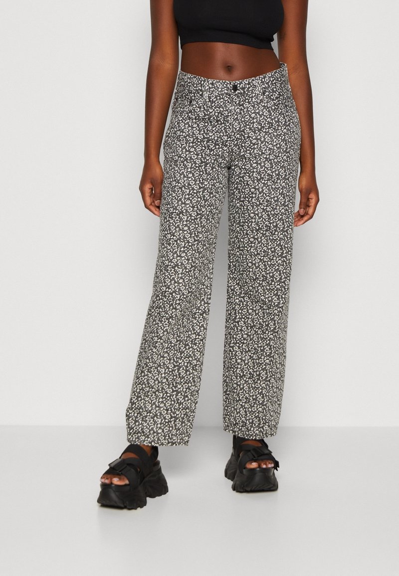 Pantalones de pierna ancha con patrón floral en blanco y negro, de cintura alta, con cierre de botón y textura de tela suave. Combinados con sandalias negras de suela gruesa.