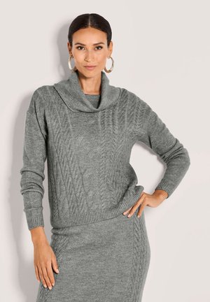 Eine Frau trägt einen grauen Kabelstrickpullover mit Schalkragen und einen passenden Rock. Sie posiert mit einer Hand auf der Hüfte vor einem einfarbigen Hintergrund.