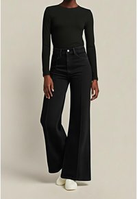 Femme portant un haut noir ajusté à manches longues, un pantalon noir taille haute à jambes larges et des chaussures blanches, debout sur un fond neutre.
