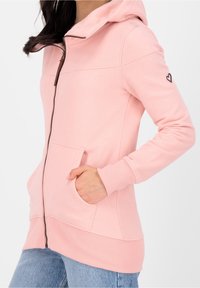 Donna che indossa una felpa leggera color rosa chiaro con zip, dotata di tasche frontali e un piccolo emblema a forma di cuore sulla manica sinistra, abbinata a jeans blu.