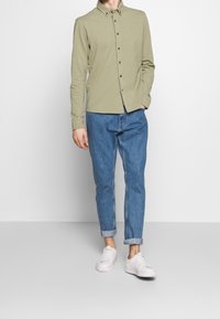 Světle olivová košile s dlouhým rukávem a límečkem na knoflíky, kombinovaná s manžetovými modrými džíny a bílými teniskami. Jednoduchý design s nenápadným brandingem.