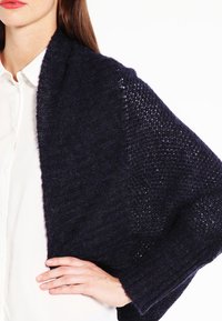 Tmavě modrý pletený šál s texturovaným vzorem, otevřenou tkaninou a hladkými okraji, přehodil přes bílou košili na knoflíky.