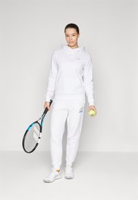 Weißer Hoodie mit Fronttasche und dezentem Logo, kombiniert mit weißen Jogginghosen mit farbigem Akzent und Markenlogo. Hält einen Tennisschläger und einen Ball.