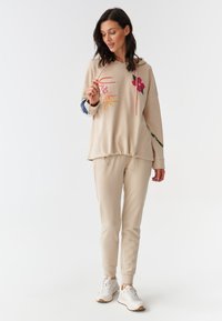 Sudadera beige con bordados coloridos, cuello con cordón y acentos florales; combinada con joggers beige a juego y zapatillas blancas.