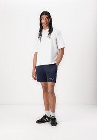 Weißes T-Shirt mit kurzen Ärmeln, kombiniert mit navyblauen Shorts mit gesticktem Grafikmotiv. Schwarze Sneaker mit weißen Akzenten und Socken in Knöchellänge.