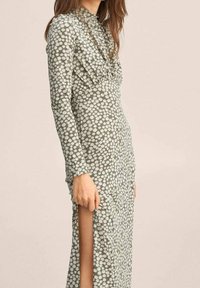 Groene bloemenjurk met lange mouwen, voorzien van een hoge ronde hals en een zijsplit. De stof heeft een zachte structuur en witte madeliefjespatronen.