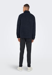 Granatowa bluza polarowa z kołnierzem, lekko teksturowana tkanina i mankiety na guziki, w połączeniu z ciemnymi spodniami i białymi trampkami.