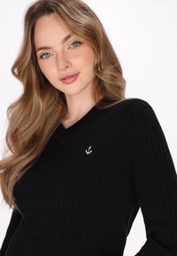 Pulover negru cu model ribbat, cu decolteu în V, având o mică broderie albă cu ancora pe partea stângă. Material moale, tricotat texturat.