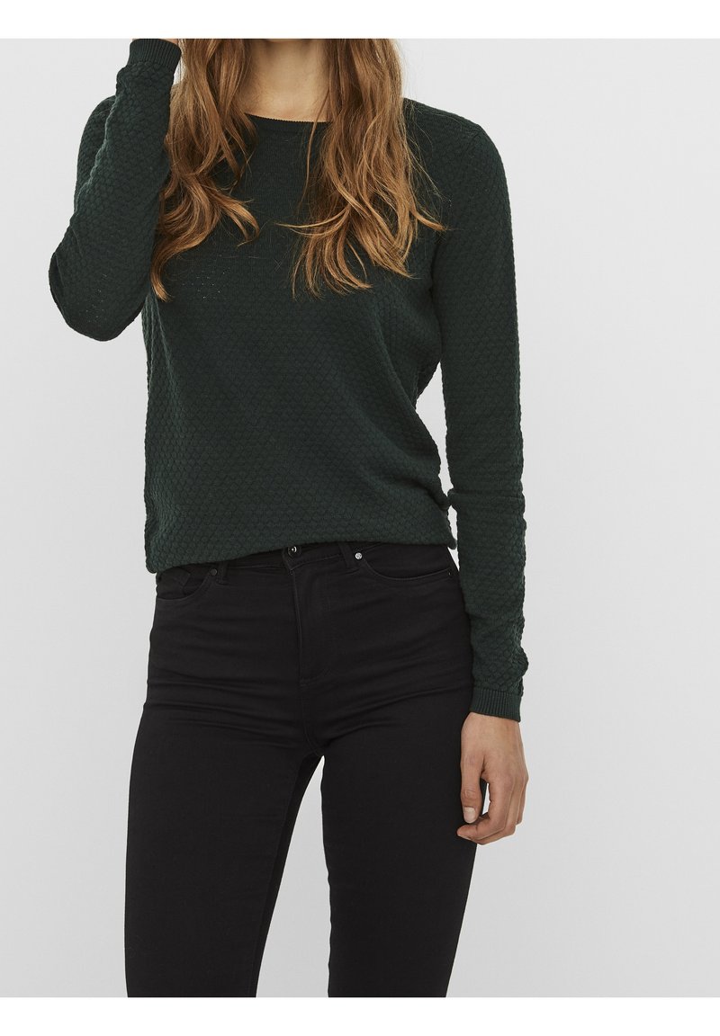 Femme portant un pull texturé vert foncé à manches longues et un pantalon taille haute noir sur un fond gris clair.