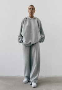 Jasnoszara, oversize bluza z kapturem i luźne spodnie dresowe, wykonane z miękkiego materiału, z kieszenią kangurką i minimalnymi szwami. Białe obuwie.