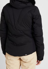 Svart puffjacka med huva och pälsdetalj, med en texturerad yta, vadderat mönster och en logotagg i den nedre delen av ryggen.