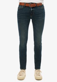 Jeans de mezclilla azul oscuro con corte ajustado, que presentan un desgaste sutil, un cinturón de cuero marrón y un diseño clásico de cinco bolsillos.