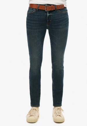 Jeans de mezclilla azul oscuro con corte ajustado, que presentan un desgaste sutil, un cinturón de cuero marrón y un diseño clásico de cinco bolsillos.