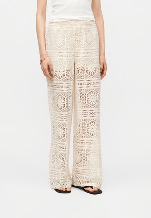 Γυναίκα που φοράει μπεζ πλεκτό παντελόνι με φαρδιά μπατζάκια και γεωμετρικό σχέδιο, καθώς και μαύρα πέδιλα με ανοιχτά δάχτυλα, στέκεται μπροστά σε λευκό φόντο.