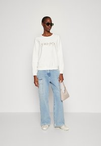 Felpa bianca con logo metallico, jeans cargo a gamba larga blu chiaro, borsa beige e occhiali da sole neri. Outfit casual con dettagli testurizzati.