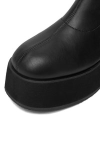 Zapato de plataforma negro de cuero con suela gruesa y costuras visibles a lo largo de los paneles frontal y lateral.