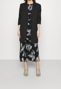 Černý blazer přes černé šaty s bílým vzorem palmových listů, doplněné zelenými pletenými podpatky. Hladká tkanina, uvolněný střih, šaty ke kolenům.