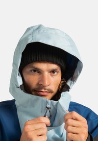 Jasnoniebieska wodoodporna kurtka z kapturem, z czarnym suwakiem i kontrastującą niebieską dolną częścią, noszona na czarnej dzianinowej czapce.