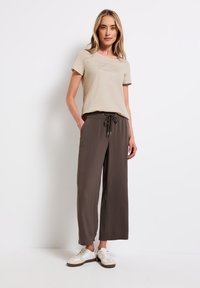 Světle béžové tričko s texturovaným designem na přední straně, spárováno s tmavě hnědými kalhotami širokého střihu, které mají elastický pas a stahovací šňůrku.