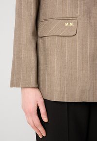 Brauner Blazer mit Nadelstreifen aus strukturiertem Stoff, versehen mit einer eingesetzten Tasche und goldenen Initialen "M.M." auf der Tasche. Schwarze Hose darunter sichtbar.