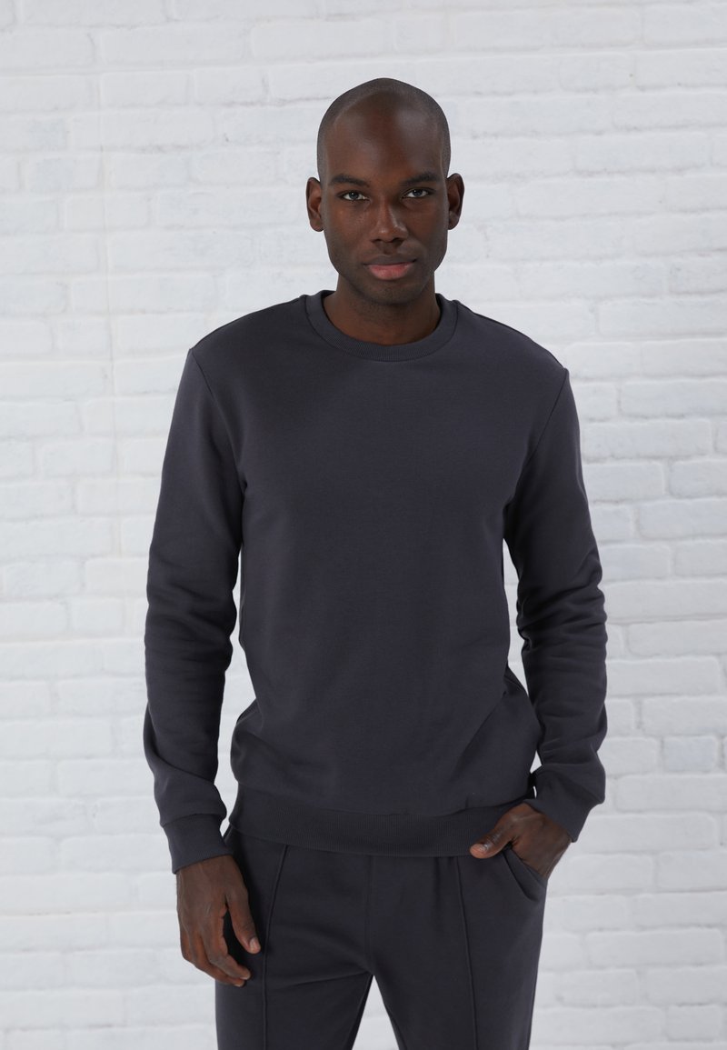 Sweatshirt gris foncé avec encolure ras du cou et manches longues, présentant une texture lisse ainsi que des poignets et un ourlet côtelés, porté avec un pantalon assorti.
