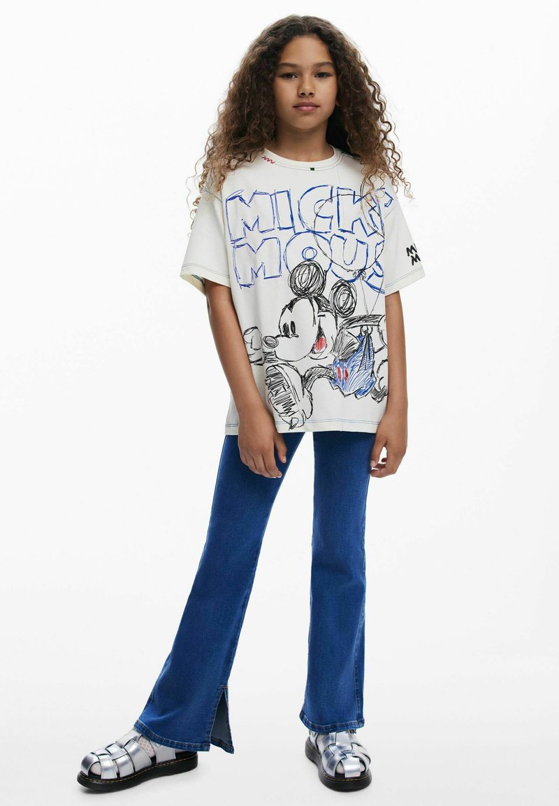 T-shirt blanc surdimensionné avec un motif coloré de Mickey Mouse, associé à un jean évasé bleu et des sandales à plateforme argentées avec des brides.