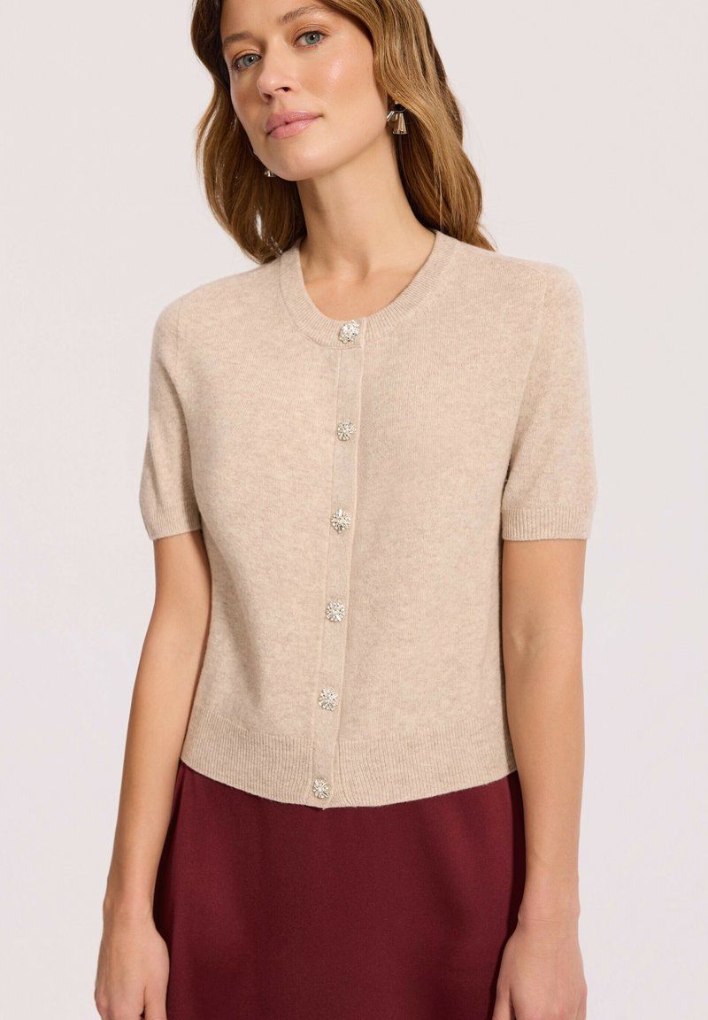 Kobieta nosząca beżowy kardigan z krótkim rękawem z ozdobnymi guzikami i bordową spódnicę, stojąca na jednolitym tle.