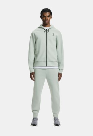 Młody mężczyzna stoi, ubrany w jasnorozowy zielony kaptur na zamek, pasujące spodnie dresowe, białą podkoszulkę i szare sportowe buty na jednolitym tle.