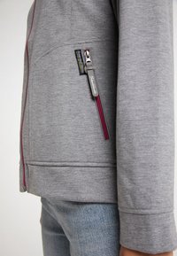 Veste grise zippée en tissu texturé, ornée d'une fermeture éclair et de détails en marron bordeaux. Comprend un petit patch logo sur le côté supérieur gauche.