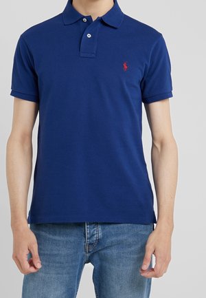 Mand, der bærer en mørkeblå kortærmet polo med en rød broderet logo og lyseblå jeans mod en grå baggrund.