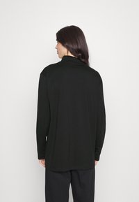 Schwarzes Langarmoberteil mit hohem Kragen, weichem Stoff, lockerer Passform und abgerundetem Saum. Getragen mit lockeren schwarzen Hosen.