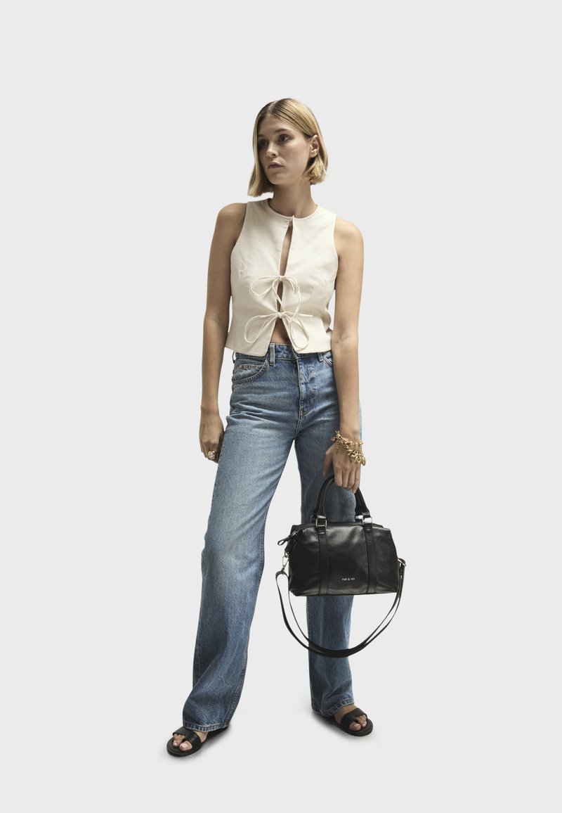 Top blanco sin mangas con un detalle de lazo central, combinado con jeans acampanados de un azul claro y un bolso de cuero negro. Texturas suaves en todo.