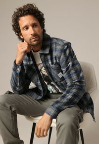 Blauwe en grijze geblokte jas met een kraag, twee zakken en een rits. Draag het over een graphic t-shirt en combineer het met grijze broek.