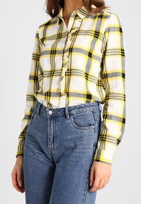 Camisa de cuadros abotonada en blanco, amarillo y gris; presenta un pliegue rizado en la tapeta delantera, mangas largas y se combina con unos vaqueros azul de cintura alta.